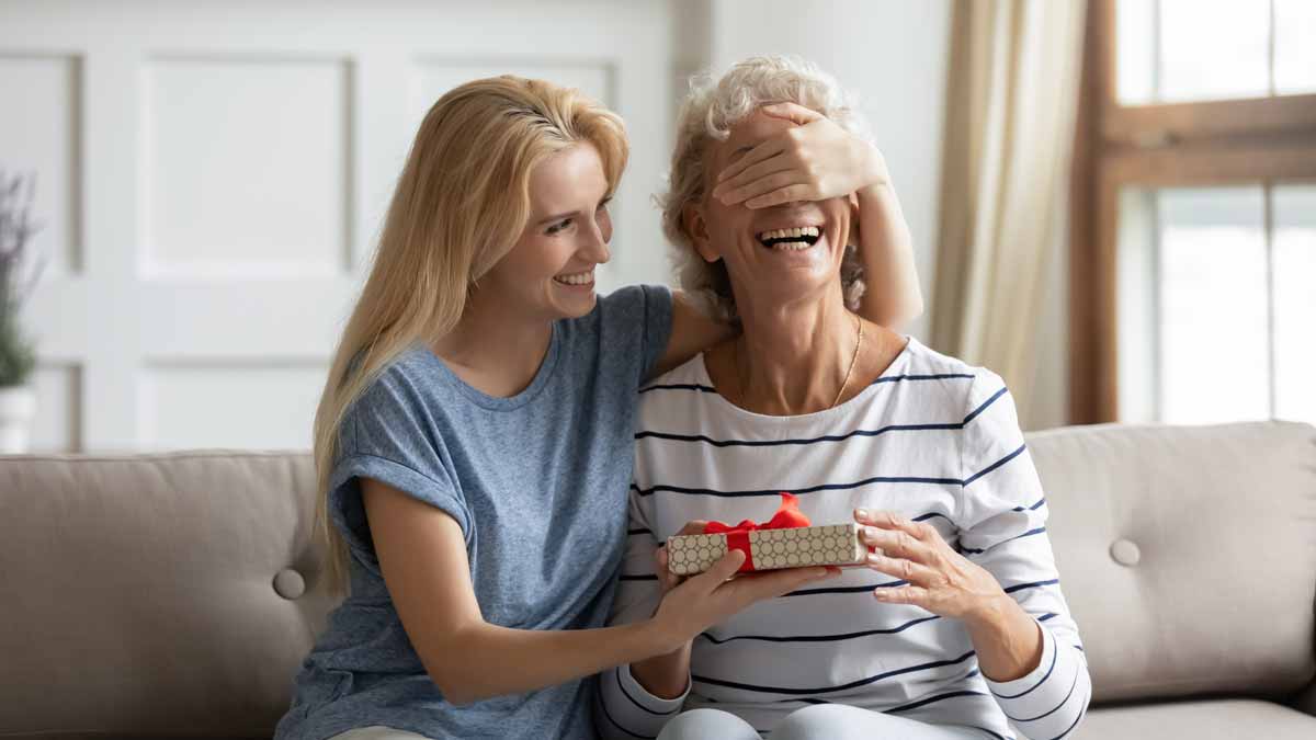 Woman covers her mother’s eyes while handing her a wrapped gift